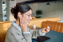 Person wearing glasses sits at a table in a cafe, placing a white wireless earbud into one ear while holding the matching earbud in the other hand. A tablet rests on the table, with seating and soft lighting visible in the background.
