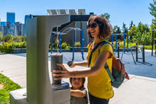 Person fills a reusable bottle at an outdoor water fountain on a sunny day while a child stands nearby in a park setting.