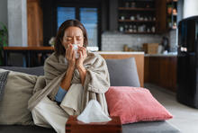 Person wrapped in a blanket sits on a couch at home holding a tissue to their nose. A tissue box rests on a table in the foreground, suggesting cold or respiratory illness symptoms.
