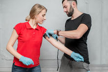 A man wearing blue gloves adjusts a blood pressure cuff on a woman’s arm as she stands in a red polo shirt against a gray wall.