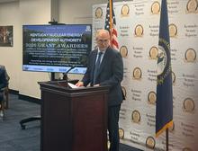 A speaker stands at a wooden podium in a government meeting room, with U.S. and Kentucky flags behind them. A screen displays “Kentucky Nuclear Energy Development Authority 2026 Grant Awardees,” and a Kentucky Senate Majority backdrop fills the wall.