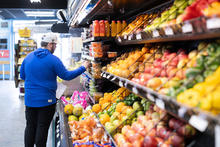 A shopper in a blue jacket and white cap browses a grocery store produce section, reaching toward packaged foods while standing beside shelves stocked with colorful fruits, vegetables and bottled juices.
