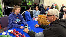 People sit around a blue-covered table at a community event, engaging in an activity with small red-and-white items and a basket on the table.