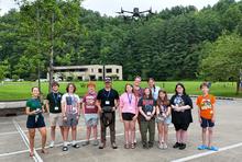 A group of students and instructors pose outdoors beneath a hovering drone. One instructor holds a drone controller while the participants stand together in a parking area surrounded by trees and a research facility.