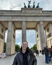 Person standing in front of the Brandenburg Gate in Berlin, Germany, with the landmark’s columns and the Quadriga sculpture visible overhead. Pedestrians and an overcast sky appear in the background.
