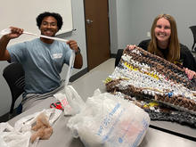 Two people sit at a table, smiling while turning plastic bags into a woven mat.