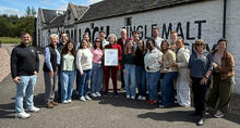 A group of about 20 people stand outdoors in front of a white stone building with large black painted lettering on the wall. One person in the center holds a framed certificate while the group gathers around for a photo.
