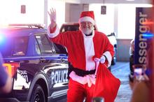 A person dressed as Santa Claus waves while walking beside a police vehicle with flashing lights, carrying a large red sack.