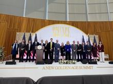 People stand on a stage beneath a large “A New Golden Age Summit” sign, with U.S. flags displayed behind them and a speaker at a podium addressing the audience.  
