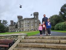 A group of four people stands on stone steps in front of a large gray stone castle with multiple towers and ornate windows. The castle is surrounded by green lawns and trees under an overcast sky. Several birds are flying above the building. 
