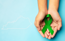 Two cupped hands hold a green awareness ribbon against a light blue background with a white outline of the state of Kentucky.