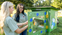 Two people standing outdoors beside a hand-painted green Little Free Library decorated with blue, white, and purple flowers. One person opens the library door while both look at the children's books displayed on the shelves inside. 