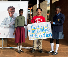 Three students hold a sign reading “Thank You UK College of Education” beside a banner at a school.