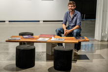 A person sits behind a low wooden bench displaying colorful books and printed materials, with two black stools in front, inside a gallery space.