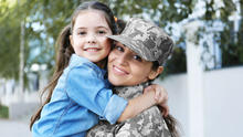 Person in military camouflage uniform embraces a young child outdoors near a building and trees.