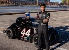 A person in a black racing fire suit stands with arms crossed in front of a black Legend race car marked with the number 44. The car features multiple sponsor logos, including the UK Alumni Association, and a colorful racing helmet rests on the roof.
