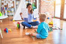An educator sits on the floor with a young child in a playroom, holding drawings of a happy and sad face while the child plays with colorful building blocks.