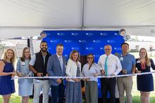 Nine people participate in a ribbon-cutting ceremony under a white tent, standing in front of a blue UK Golisano Children’s Network backdrop and holding gold ceremonial scissors as they cut a white ribbon.