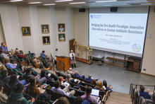Presenter stands at the front of a lecture hall, speaking to an audience seated in tiered rows. A large projection screen displays a presentation titled “Bridging the One Health Paradigm.”
