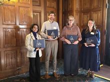 Four people stand side by side indoors in front of ornate wooden doors, each holding a plaque and smiling at the camera during UK Staff Senate awards presentation.