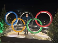 A man stands inside the Olympic rings display at night with snowy mountains in the background.