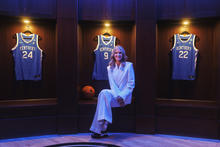 A person dressed in a light-colored suit sits in the center of a display featuring framed University of Kentucky basketball jerseys. Three blue jerseys, including numbers 24, 3 and 22, are mounted in illuminated wooden display cases behind the person.