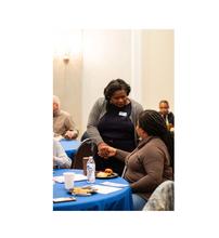 Two attendees greet each other with a handshake during a networking or community event. The interaction takes place at a round table covered with a blue tablecloth, with notepads, a water bottle, and breakfast items visible. 
