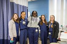 Five students stand together in a clinical setting, with exam room curtains and medical equipment visible in the background.