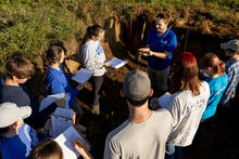 Group of students standing around a soil pit outdoors while an instructor holds up a sample of soil, explaining its characteristics. Several students take notes on clipboards, and tools are visible in the exposed layers of earth behind them.