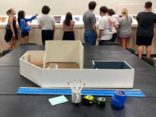 Model of an art gallery sitting on a table with a ruler, a cup of pens, tape and a tape measure, with students standing in the background looking at an exhibit with their backs to the camera