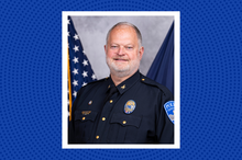 Police chief in a dark police uniform poses for a professional headshot. An American flag and a Kentucky state flag are visible in the background, with a blue patterned graphic border framing the portrait.