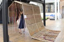 A person walks through a large, tent-like textile installation made of woven fabric panels and hanging threads, set up inside an open, light-filled gallery space with wooden floors.