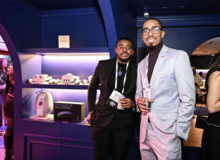 Two men wearing conference badges pose and smile at a networking event inside a modern, blue-lit venue. One wears a black suit and holds a drink, while the other wears a light gray suit with a dark turtleneck and glasses.
