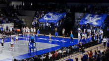  Fans hold large blue Kentucky Wildcats flags in the stands as UK volleyball players stand on the court.
