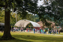 A group of children and adults walk along a gravel path at an outdoor agricultural facility, gathering near livestock pens and a covered barn structure. Large shade trees frame the scene in the foreground.