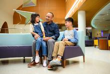 Family sits together on a curved bench in a bright hospital lobby, with one child seated on an adult’s lap and another child beside them. Modern star-shaped hanging art and warm wood accents are visible in the background.