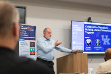 Chief Monroe leads a session during the Safer Kentucky Symposium, with a screen displaying “Collaborative Discussion: Building a Safer Commonwealth Together” and event sponsor branding.