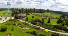 Aerial view of a landscaped green space with winding paths, gardens, trees and a building, with a water tower visible in the distance.