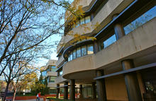 Exterior view of a multi-story campus building with curved concrete balconies, large windows and cylindrical support columns. Bare tree branches frame the left side of the image, while a sidewalk and landscaped areas surround the building.