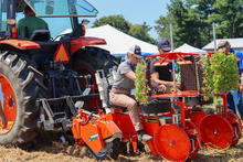 A large orange tractor is attached to a red vegetable transplanter during an outdoor agricultural equipment demonstration. Several people are gathered around the machine, with one person seated at the planting station loading seedling trays.