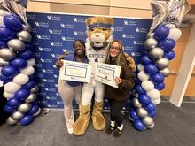 Two people pose with the UK Wildcat mascot in front of a College of Education backdrop, framed by blue, white and silver balloons. They hold signs reading “The heart to be the next Elementary Teacher” and “The heart to be the next Wildcat.”