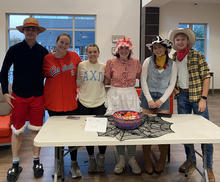 Six people, several wearing costumes, pose behind a table with a bowl of candy and a spiderweb decoration.