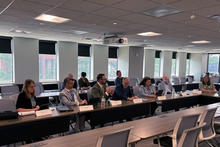 A group of people seated around U-shaped conference tables during a meeting in a large room with windows, laptops, notebooks, water bottles and name placards arranged on the tables.
