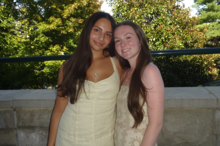 Two people stand side by side outdoors in front of a stone railing, with trees and greenery in the background. One person wears a light-colored sleeveless dress, and the other wears a light pink long-sleeve top with a light-colored skirt.