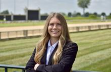 A person with long hair stands outdoors with arms crossed, wearing a dark blazer over a white shirt, with a grassy field and railing in the background.