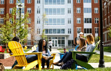 Students sit in colorful Adirondack chairs on a campus lawn, gathered in a circle and talking outside a residence hall.