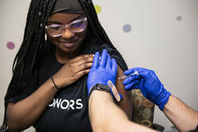 A health care worker wearing blue gloves administers a vaccine injection into a person's upper arm while the person holds up a shirt sleeve.