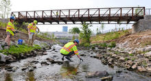 Three researchers in high-visibility safety vests and hard hats collect water samples in a shallow rocky stream beneath a metal pedestrian bridge, with stabilized creek banks and a commercial building visible in the background.