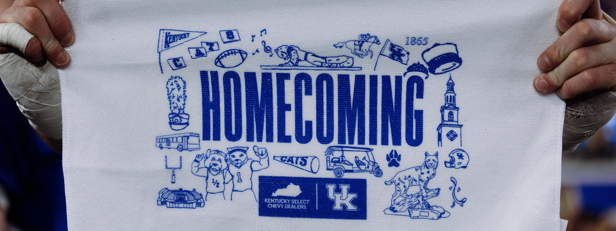 Close-up of a person holding a white rally towel featuring a blue “Homecoming” design with University of Kentucky branding and illustrated campus-themed icons. A blurred crowd and another person are visible in the background.