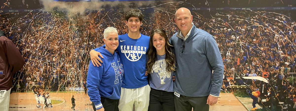 Four people pose together in front of a large mural depicting a University of Kentucky basketball championship celebration. The group stands with their arms around one another, wearing Kentucky-themed clothing, confetti and a packed arena crowd in backgro